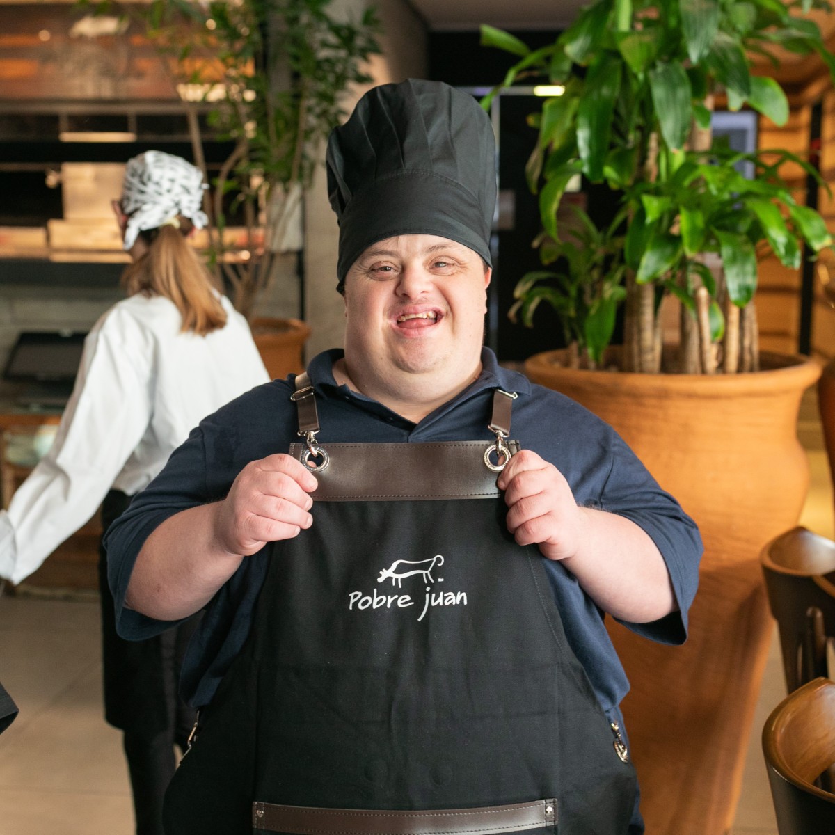 Jovem com síndrome de Down, de touca e avental de cozinha do restaurante Pobre Juan, sorrindo no salão.